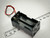 A 4 AA battery holder with black plastic casing and metal spring terminals, accompanied by red and black leads for connections. The holder is positioned beside a ruler marked in millimeters. Available at PartsMine.com.