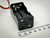 Vertical image of a 4 AA battery holder with metal spring terminals and red-black leads, standing on a surface with a millimeter ruler beside it. Available at PartsMine.com.
