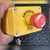 Hand holding yellow emergency stop enclosure, close view of red mushroom operator and EMERGENCY STOP legend