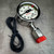 Angled view of the polished-metal pressure switch gauge, coiled black lead, red cap