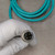 M12 D-code male connector face-on; four gold pins clean and intact, knurled metal coupling ring with light surface oxidation. Teal jacket coiled, no kinks visible.