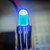 Blue LED illuminated, emitting cyan-blue light through clear 5mm-style dome. Both leads straight and untrimmed with latch tabs intact.