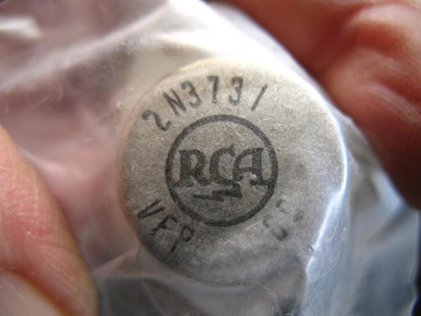 Closeup image of an RCA 2N3731 transistor, clearly displaying the circular RCA logo stamped on its metallic gray top. The model number 2N3731 is visible in an arc above the logo, with additional manufacturing codes like VFP partially seen below. The electronic component appears to be in clear protective packaging, held by fingers, suggesting a new old stock (NOS) condition or a carefully presented vintage part. This specific semiconductor is a soughtafter power transistor for electronics repair, audio amplifier projects, or as a replacement part for industrial and consumer devices.