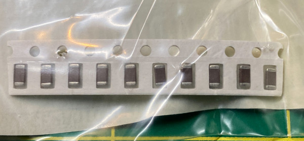 Ten dark ceramic chip capacitors on paper tape inside a bag, green mat behind