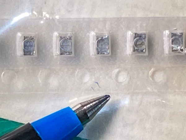 Five very small gray chip capacitors in clear carrier tape near pen tip