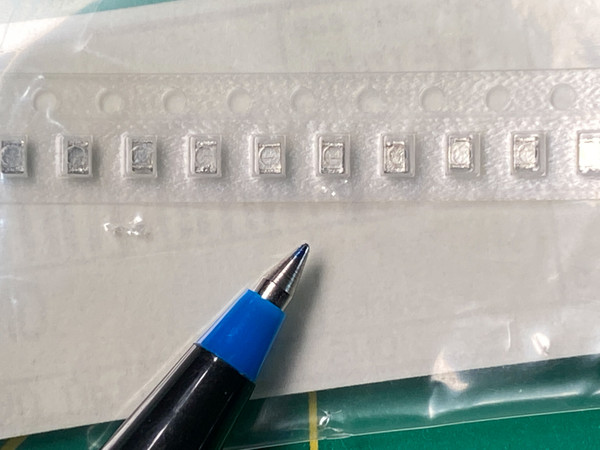 Row of ten gray film chip capacitors in clear tape, pen tip pointing up