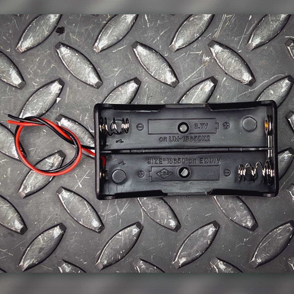 Black plastic body molded with 3.7V, UM-18650X2, SIZE 18650 OR EQUIV markings. Two spring contacts visible. Red and black wire leads attached.