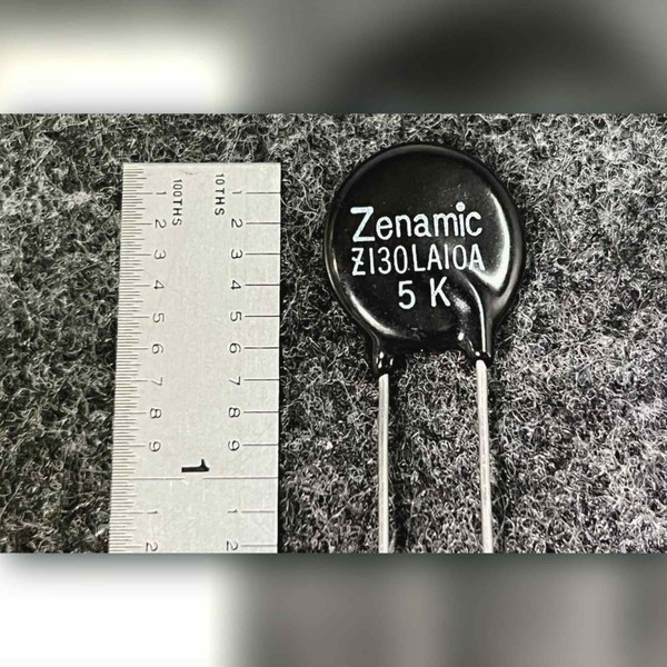 Black disc varistor printed Zenamic Z130LA10A 5K beside a vertical inch rule