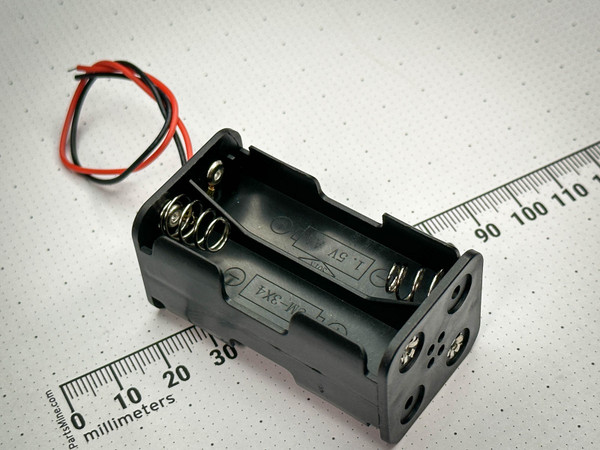 A 4 AA battery holder with black plastic casing and metal spring terminals, accompanied by red and black leads for connections. The holder is positioned beside a ruler marked in millimeters. Available at PartsMine.com.