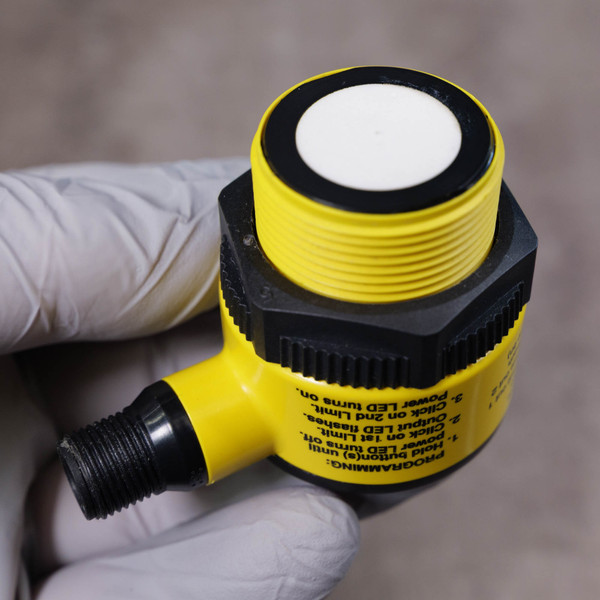 Sensor showing clean yellow housing with black nuts. M12 connector threads intact. Cracked cover with missing piece on sensor face. Programming instructions visible: "PROGRAMMING: 1. Hold button(s) until power LED turns off. 2. Click on 1st Limit. Output LED flashes. Click on 2nd Limit. 3. power LED turns on.".