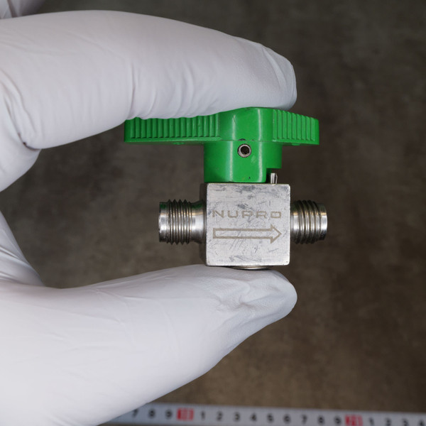 Nupro plug valve showing "NUPRO" and flow arrow. Stainless steel body has minor scuffs. Green handle shows light wear. NPT threads appear clean and intact.