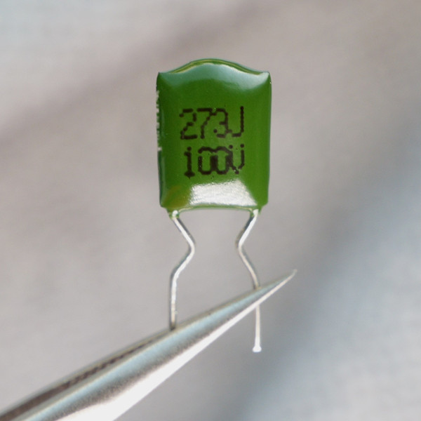 Green radial capacitor marked 273J 100V held in tweezers.