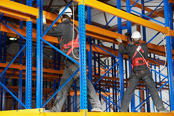 Men wearing workwear and safety hats assembling pallet racks on a raised structure, demonstrating safe warehouse construction practices.