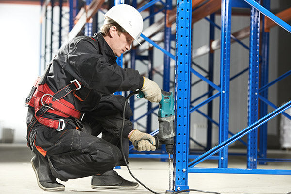 Image showing a man wearing safety gear, kneeling down alongside a partially assembled rack, carefully drilling bolts into a cement floor for a secure attachment.