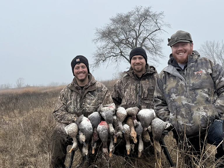 Three hunters in camo kneeling in a foggy Missouri marsh behind a row of harvested ducks after a morning waterfowl hunt