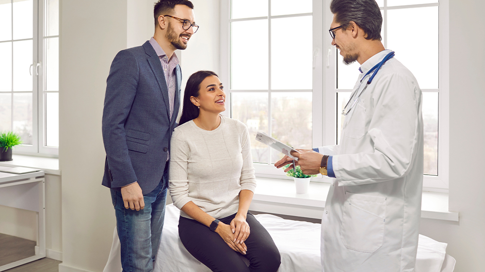 A happy couple in a window-lined exam room discussing causes of secondary infertility with their male physician.