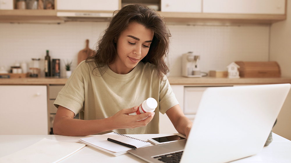 With her laptop and notepad, a young woman sits in a kitchen, reading a bottle label to learn more about Vitex for fertility.