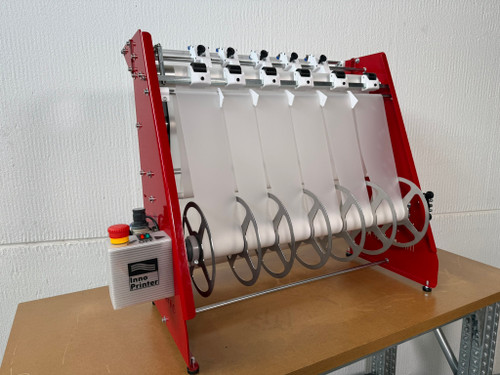 InnoPrinter RC60 DTF roll slitter threaded with white film, angled view. Red-framed slitter with six strips of white DTF film fed from the top rail down through the blade assembly and separated onto metal discs along the rewind shaft, with the InnoPrinter control box and emergency stop visible on the left.