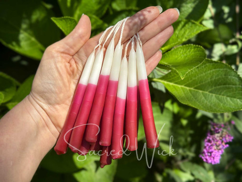 Hand holds several pairs of white and magenta ombré chime candles looped over a finger, glowing in dappled sunlight with lush greenery in the background.