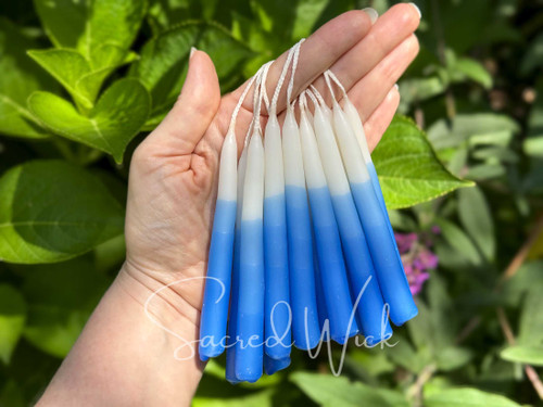 Several pairs of white and blue ombré chime candles looped over a finger, glowing in dappled sunlight with vibrant greenery in the background, showcasing the beauty of these hand-dipped beeswax candles.