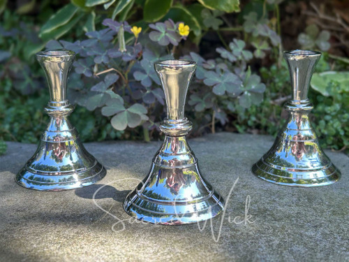 Three silver-tone brass candle holders in a vintage candle stick style with clover and greenery in the background sitting on a concrete pathway zoomed in slightly to see detail.