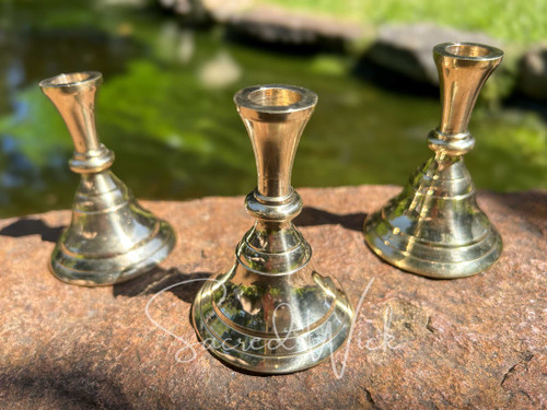 Three brass candle holders sit on brickwork in a vintage candlestick style with greenery and water behind them.
