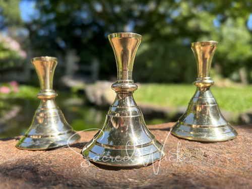 Three brass candle holders sit on brickwork in a vintage candlestick style with greenery and water behind them, shown in a close-up.