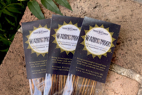 Three packs of waning moon incense sticks lie on brickwork in filtered sunlight with greenery nearby.