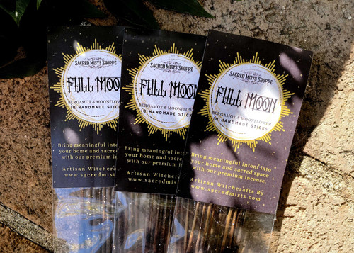 Three packs of full moon incense sticks lie on brickwork in filtered sunlight with greenery nearby.