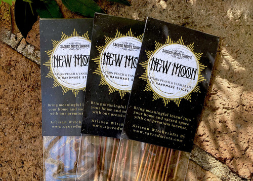 Three packs of new moon incense sticks lie on brickwork in filtered sunlight with greenery nearby.