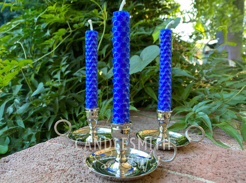 Three silver-tone chamberstick candle holders with beeswax candles are on a brick ledge, with greenery in the background.