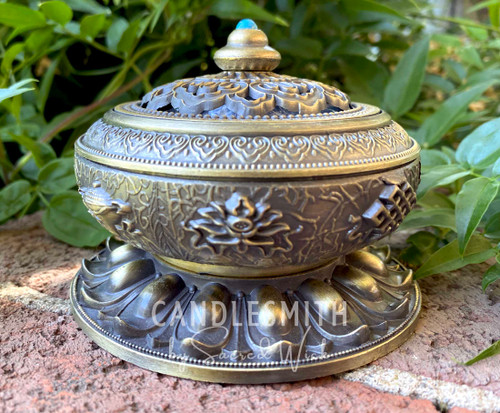 Tibetan-Style Brass Incense Bowl with Decorative Lid sits on a brick ledge with greenery on the background and shown from the front to display the markings and style.