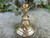A brass candle holder sits on a concrete pathway, showing its vintage styling with clover and greenery behind it shown from above slightly.
