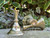 Three brass candle holders sit on a concrete pathway with clover and greenery in the rear; two candle holders are shown lying on their sides.