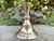 A brass candle holder sits on a concrete pathway, showing its vintage styling with clover and greenery behind it.