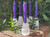 Several selenite crystal candle holders with purple beeswax candles displayed on brickwork with greenery in the background, candles not included