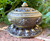 Tibetan-Style Brass Incense Bowl with Decorative Lid sits on a brick ledge with greenery on the background and shown from the front to display the markings and style.