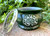 one tree of life green soapstone incense bowl with screen propped on the left side rests on brickwork with greenery int he background, shown from the front to display the tree of life design.