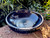 One Black Stone Shallow Incense & Candle Bowl with Polished Finish shown from slightly above rests on brickwork with grenenery in the background.