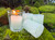 several sage bundle handmade votive candles sit on moss covered brick, with one to the side in a clear votive cup while lit displaying the candle beauty.