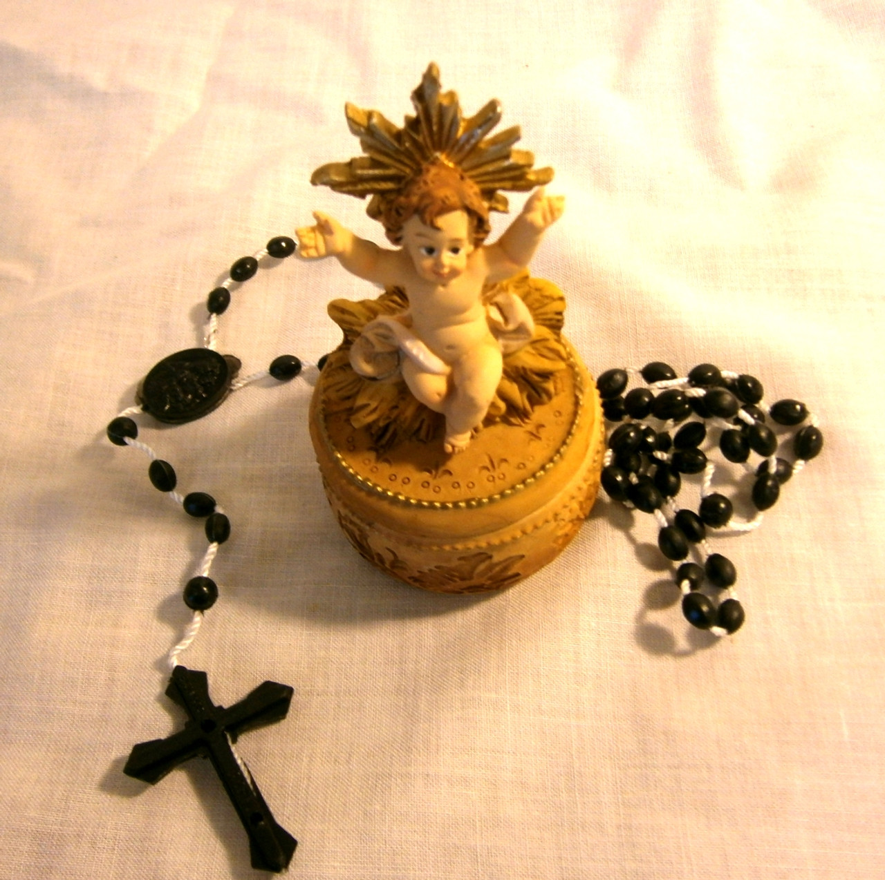 Infant Christ seated on a small, circular box containing a black plastic rosary.