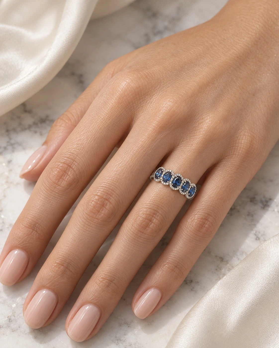 Hand wearing a five-stone royal blue sapphire and diamond halo ring in 18K white gold on a marble surface
