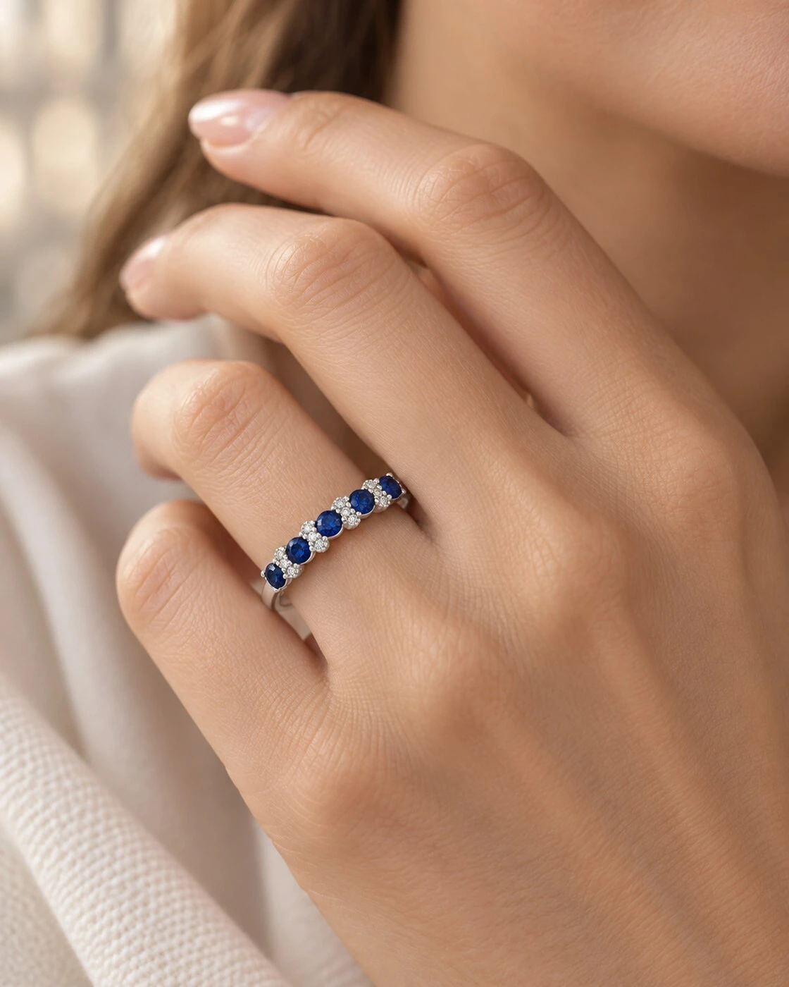 Hand wearing a royal blue sapphire and diamond stackable band ring in 18K white gold against white silk