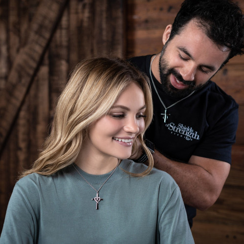 Woman wearing Shields Of Strength Women's Stainless Steel Cross Necklace accented with a Swarovski Crystal Heart-Isaiah 46:4.   Man holding her hair back to highlight the beauty of the necklace.
