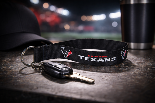 Houston Texans lanyard keychain with attached car key resting on a textured surface, set against a blurred stadium-style background with soft lighting and neutral accessories.