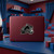 A close-up shot of a maroon laptop with a silver Colorado Avalanche logo, centered on a blue desk. The room has a maroon and blue theme, including curtains, bedding, and shelves.