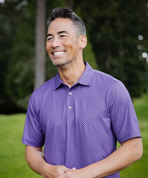 Smiling man standing on a golf course wearing a purple patterned golf polo shirt.
