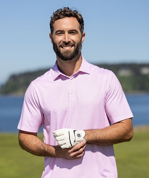 Male golfer smiling outdoors on a golf course near the water, wearing a light pink golf polo and a white golf glove.