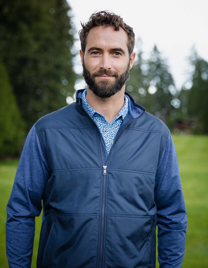Bearded man standing on a golf course wearing a blue lightweight zip-up jacket layered over a patterned collared shirt.