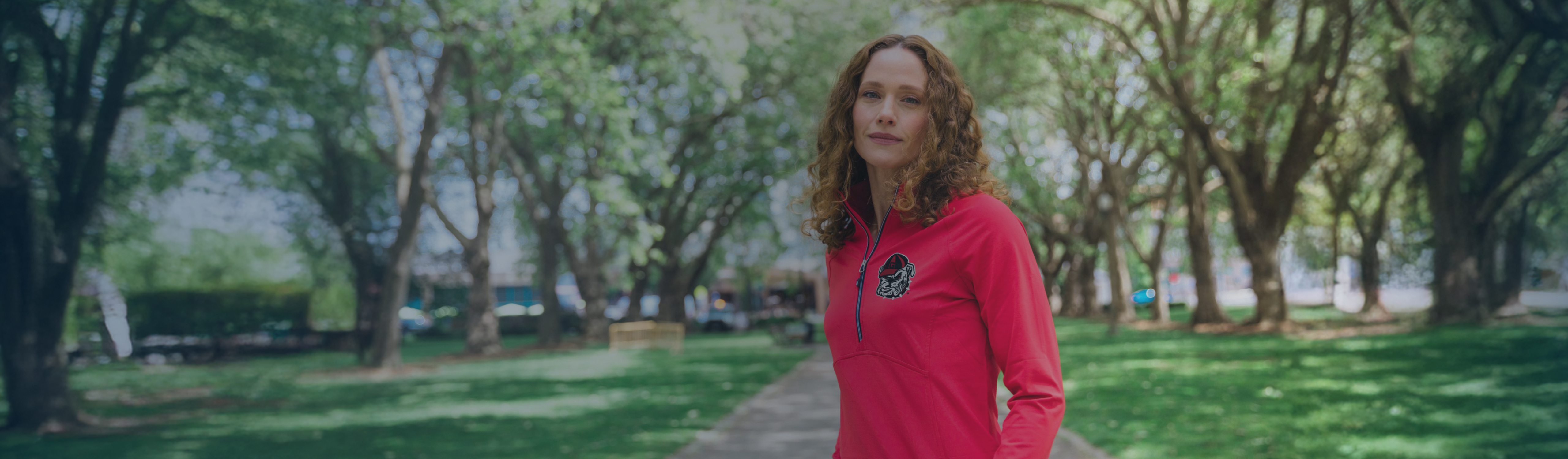 Woman wearing a bright red quarter-zip with a University of Georgia logo, standing on a tree-lined path in a sunny park.
